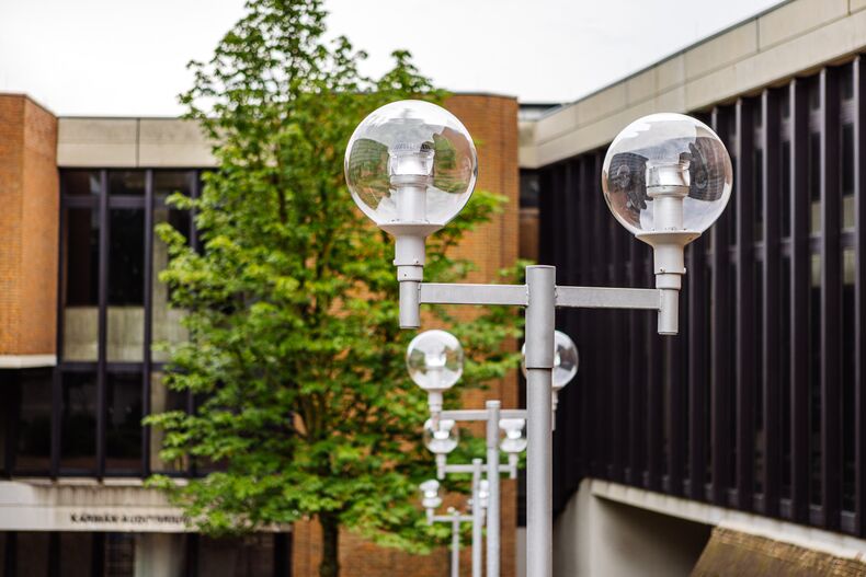 The photo shows the street lamps starting from Platanenplatz opposite the main building of RWTH Aachen University and leading down to the Karmanauditorium. In the center of the photo, a T-shaped streetlight is shown with two spherical lamps on the top right and left. The light sources in the lamps are LEDs. In the background you can see three more street lamps as well as the Karman Auditorium and a tree.