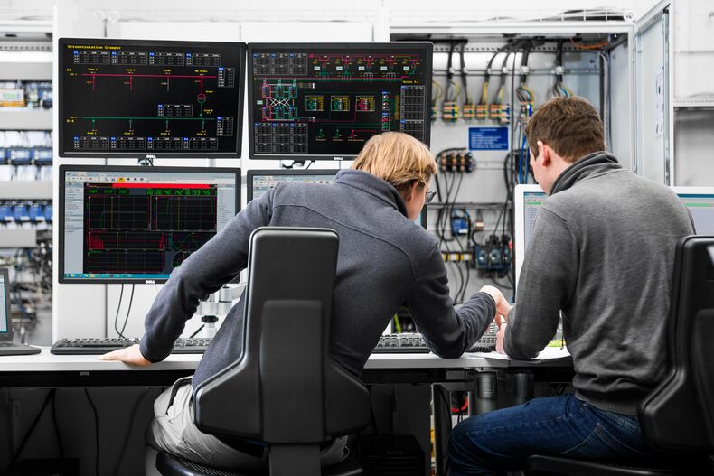 Two male persons are photographed. They are sitting at a desk with their backs to the camera. In front of them are monitors with technical data. Switching circles can be seen in the background.