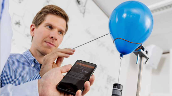 Eine Person, die mit einem langen Stab in einen Blauen Luftballon sticht. Davor ein Arm mit einem Smartphone in der Hand.