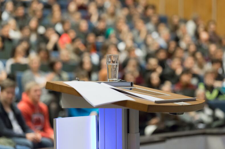 A photo of a crowded lecture hall in the foreground a lectern with slips of paper. The photo is blurred in the background.