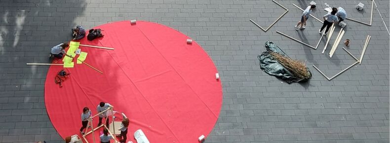 The picture shows a bird's eye view. Participants of Summer School 2022 set up art installations on a large, round red carpet and are filmed doing so.
