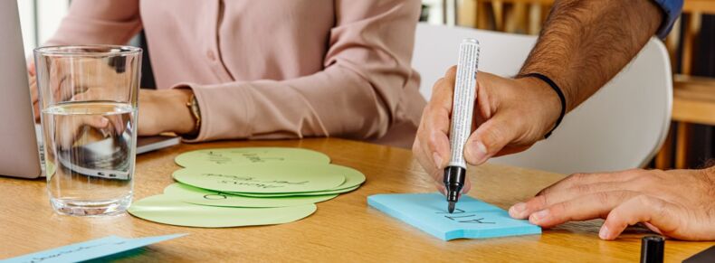 The picture shows a hand writing a note on a notepad with a pen. In the background, a person is sitting and writing on a computer.