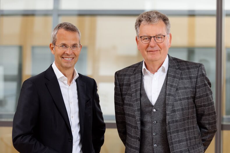 Prorectors Professor Malte Brettel, Prorector for Business and Industry, and Professor Matthias Wessling, Prorector for Research and Structure, stand in front of a glass wall.  Professor Brettel is wearing a black suit with a white shirt, Professor Wessling a gray cape jacket, gray vest and pants, and a white shirt.  They are smiling into the camera.