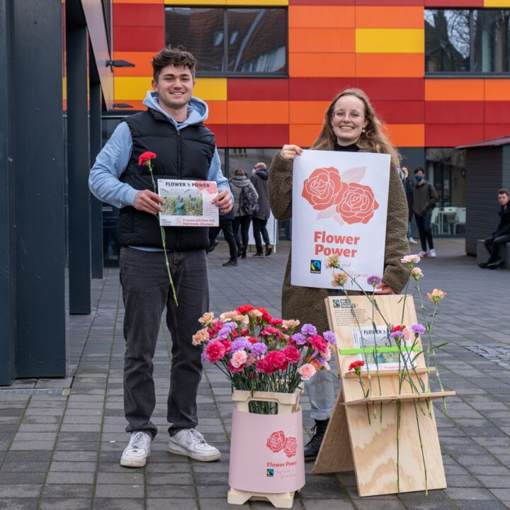 Zwei Studierende stehen vor der Mensa Academica und lächeln in die Kamera. Die linke Person hält eine Blume und ein DIN A4 großes Informationsblatt zu der Aktion Flower power in der Hand. Die rechte Person hält ein größeres Plakat mit zwei Rosen und dem Schriftzug Flower Power in den Händen. Im Vordergrund sind zwei Halterungen aus Holz abgebildet, in denen viele bunte Blumen stehen. Darunter rote, lila und rosa-beige Blüten.