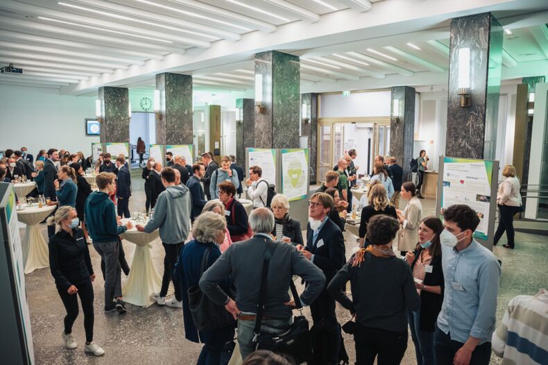 The picture shows the kick-off event of the Humboldt N initiative. Many people are standing in a large hall and talking to each other. Posters are scattered around the room.