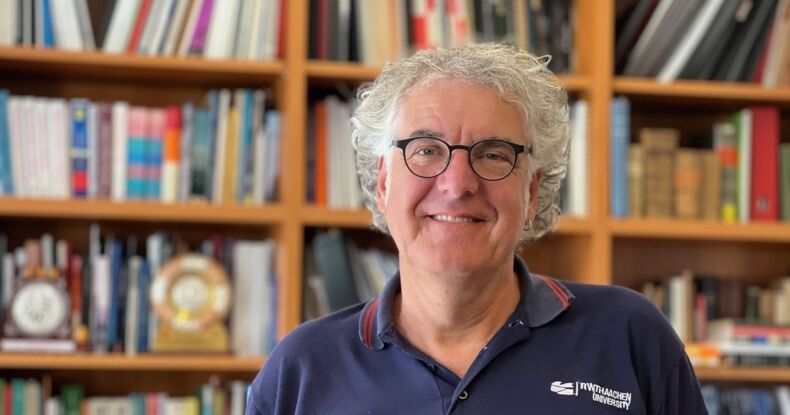 The picture shows Professor Dr. Reicherter. He is wearing a blue shirt with the inscription RWTH Aachen University. A bookshelf can be seen in the background.