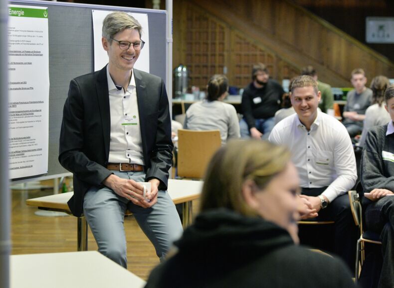 One person is sitting on a table. Other people are sitting on chairs and talking to each other. There is a poster board with posters in the background.