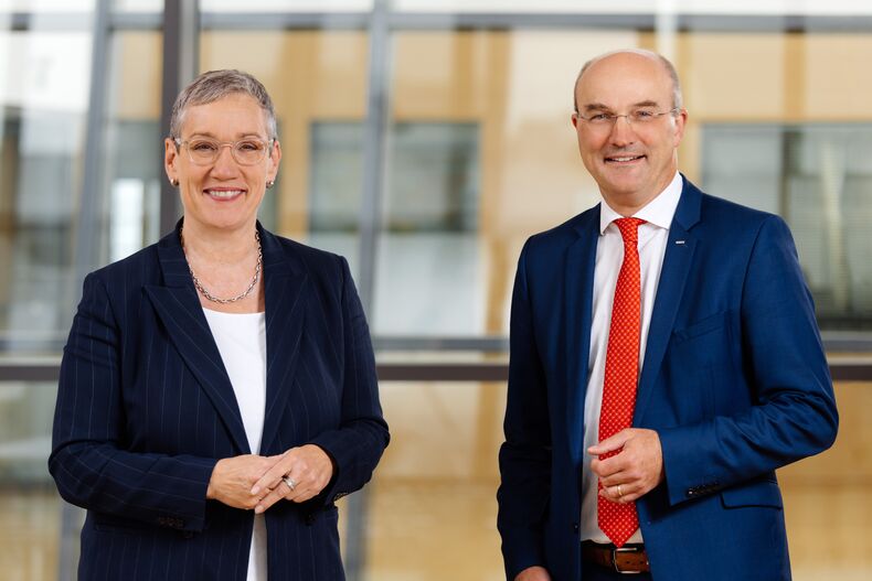 Mayor Sibylle Keupen and Rector Ulrich Rüdiger in front of a glass wall. Ms. Keupen in a dark blue blazer and white top, Professor Rüdiger in a blue suit, white shirt and red tie.  Both are smiling into the camera.