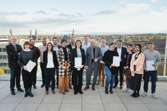 17 people, including Rector Rüdiger and the representatives of the funded projects, smile into the camera. Aachen's city center with the cathedral and town hall are in the background.