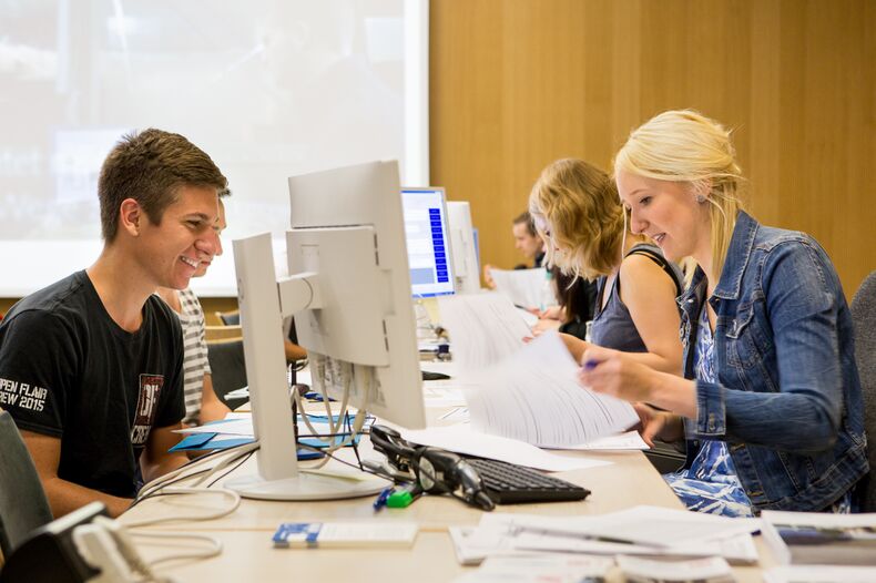 Several people at a table. In the foreground on the right a female person checking documents, in front of her on the table a computer. On the left, a male person sits in front of the table and waits. In the background a similar situation.