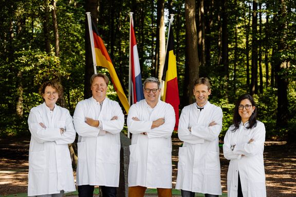 Two female and three male persons in white lab coats are standing in front of the flags of Germany, the Netherlands and Belgium at the border triangle. They have their arms crossed and are smiling at the camera.