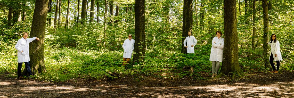 Two female and three male persons in white lab coats stand in different poses leaning against trees spread out in the forest. They smile into the camera.