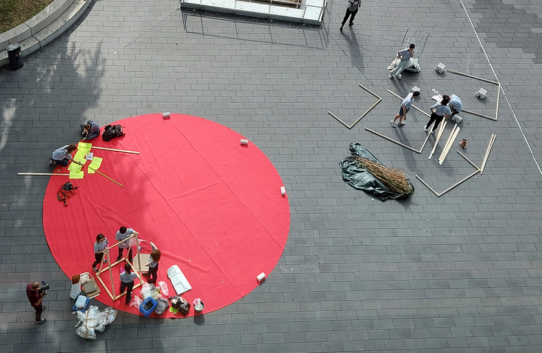 The picture shows a bird's eye view. Participants of Summer School 2022 set up art installations on a large, round red carpet and are filmed doing so.