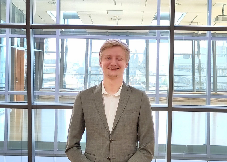 AStA Sustainability & Student Engagement Officer Florian Winkler stands in front of a glass wall. He is wearing a gray suit and a white shirt. He smiles into the camera.