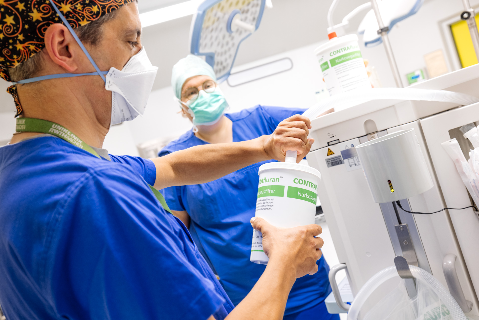 A female and a male person are standing in an operating room. They are wearing blue surgical gowns, protective medical masks and hoods. In the foreground, the male person connects a cylindrical container with anesthetic drugs to a machine with a hose.