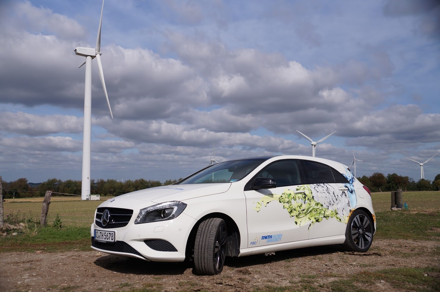 A stationary car parked in front of a field with wind turbines.