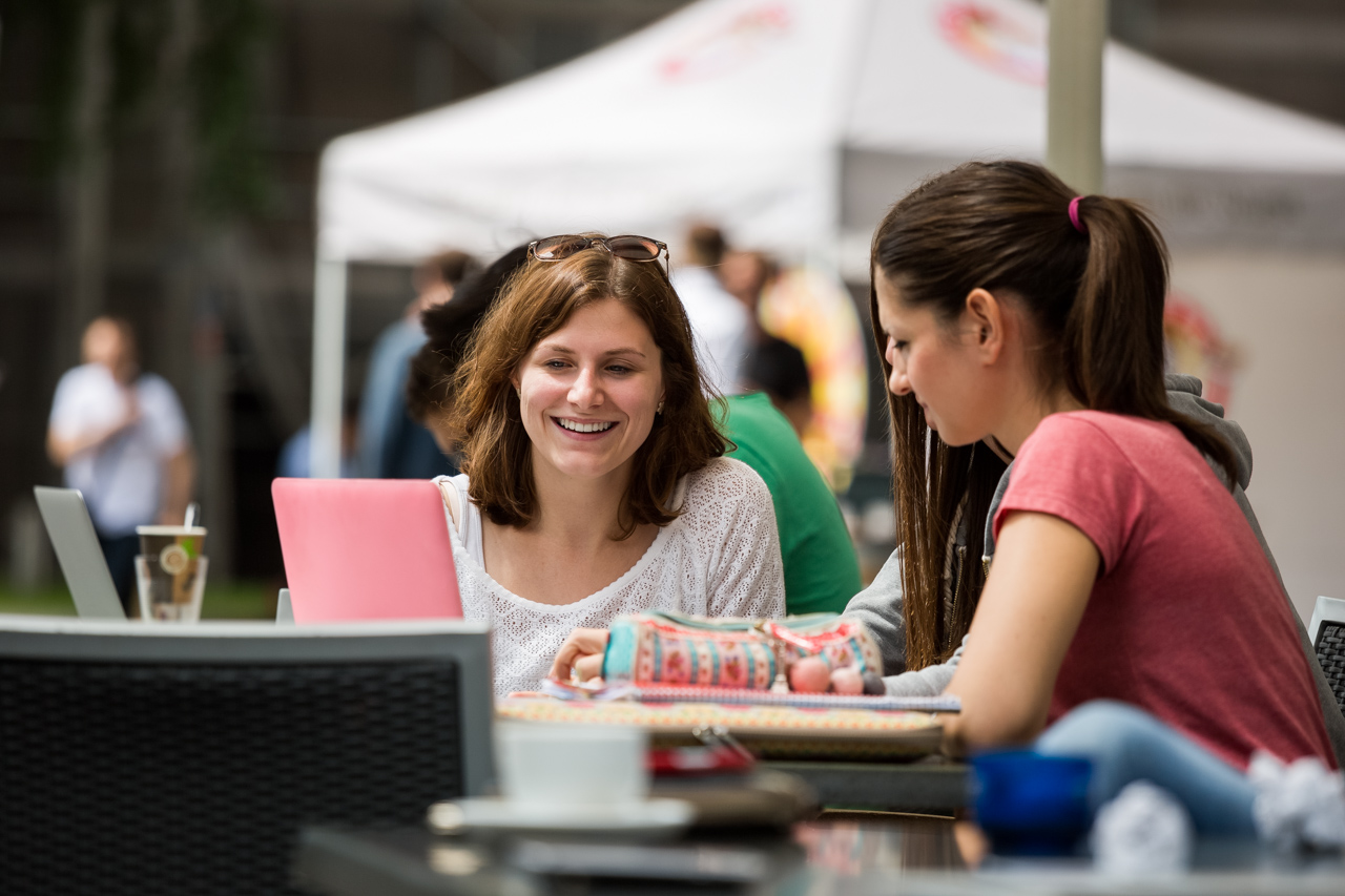 Drei weibliche Personen sitzen an einem Tisch im Freien. Darauf sind Schreibmaterialien und ein Laptop.